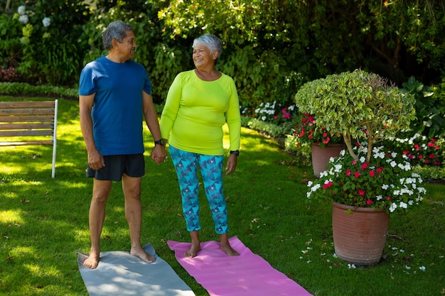 Glimlachend biraciaal senior paar hand in hand terwijl ze op yogamatten staan tegen planten in de tuin