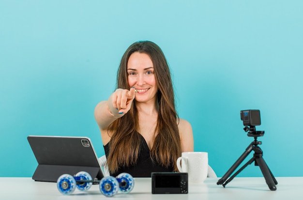 Glimlachend bloggermeisje kijkt naar de camera door de camera met wijsvinger op een blauwe achtergrond te richten