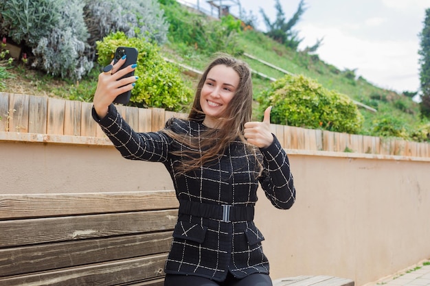 Glimlachend bloggermeisje neemt selfie met haar smartphone door een perfect gebaar in het park te tonen