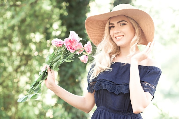 Glimlachend blond meisje dat bloemen vasthoudt met een strohoed in de openlucht Kijkend naar de camera Zomerseizoen