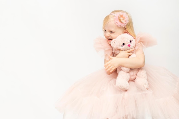 Glimlachend blond meisje in weelderige roze jurk en hoepel decoratie Witte achtergrond Gelukkig klein meisje hugs teddybeer