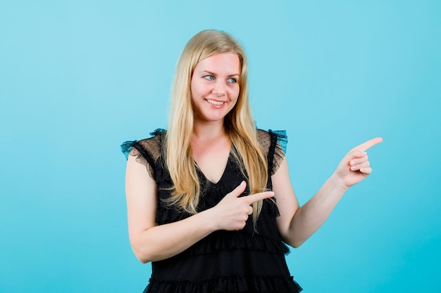 Glimlachend blond meisje kijkt en wijst naar rechts met wijsvingers op blauwe achtergrond