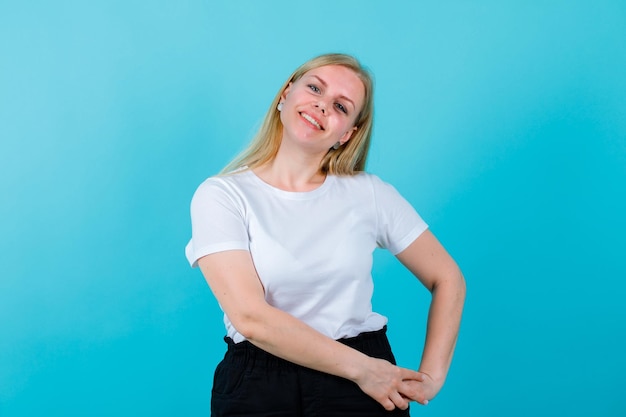 Glimlachend blond meisje kijkt naar de camera door de handen op de rechterkant van de taille op een blauwe achtergrond te leggen