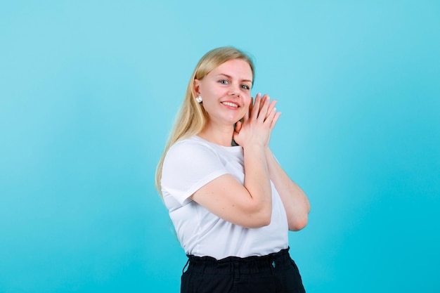 Glimlachend blond meisje kijkt naar de camera door handen bij elkaar te houden op de schouder op een blauwe achtergrond