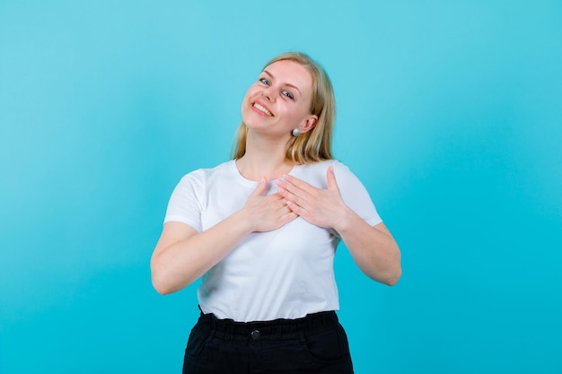 Glimlachend blond meisje kijkt weg door handen op de borst te houden op een blauwe achtergrond