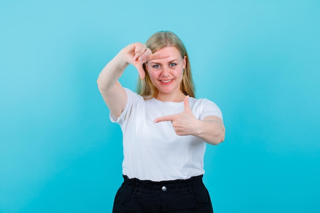 Glimlachend blond meisje zaait framegebaar op blauwe achtergrond