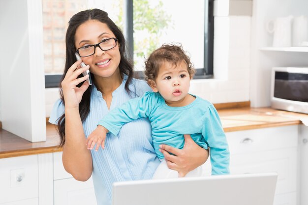 Glimlachend brunette die haar baby houden en laptop op telefoongesprek in de keuken met behulp van