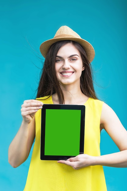 Glimlachend brunette meisje gekleed in geel shirt en hoed houdt tabletcomputer met groen scherm vast voordat blauwe achtergrond ruimteconcept adverteert