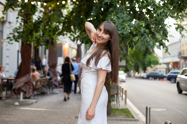 Glimlachend brunette meisje met lang haar in witte jurk op een achtergrond van groene bomen in de stad