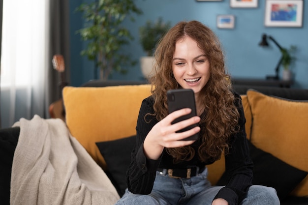 Glimlachend brunette tienermeisje met krullend haar gebruikt smartphone terwijl ze op de bank zit