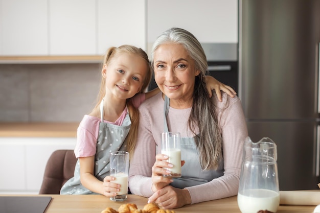 Glimlachend europees oud vrouwtje en klein meisje in schorten met glazen melk genieten van croissants in keukeninterieur