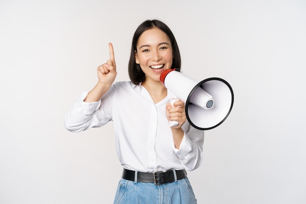 Glimlachend gelukkig Aziatisch meisje dat in een megafoon praat en omhoog wijst en kortingspromo aankondigt met advertentie bovenop die op een witte achtergrond staat