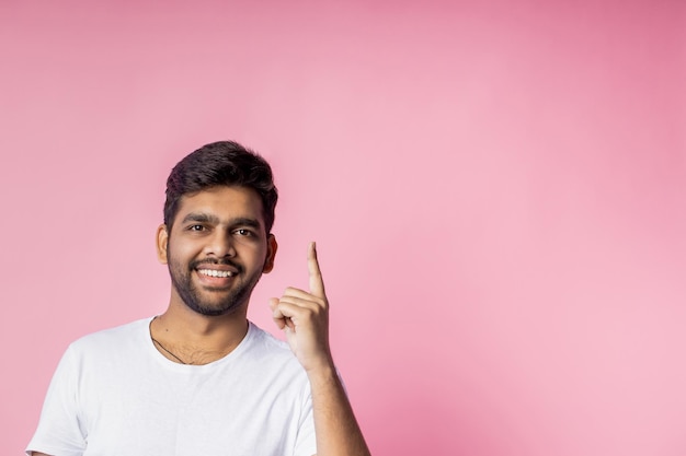 Glimlachend gelukkig bebaarde jonge man die naar boven wijst met wijsvinger, gekleed in wit casual t-shirt, reclame voor nieuw item, geïsoleerd op roze achtergrond. advertentie, gebaar concept.