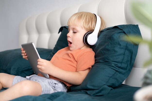 Glimlachend gelukkig jongetje met tablet in draadloze koptelefoon in bed thuis.