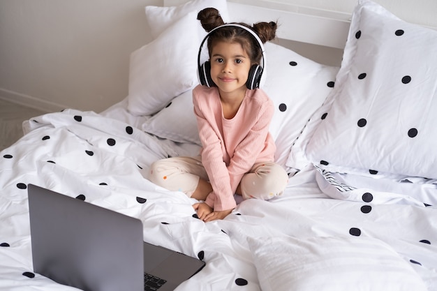 Glimlachend gelukkig klein meisje met laptop in draadloze koptelefoon in bed thuis