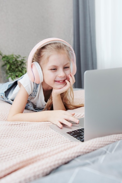 Glimlachend gelukkig klein meisje met laptop in draadloze koptelefoon in bed thuis