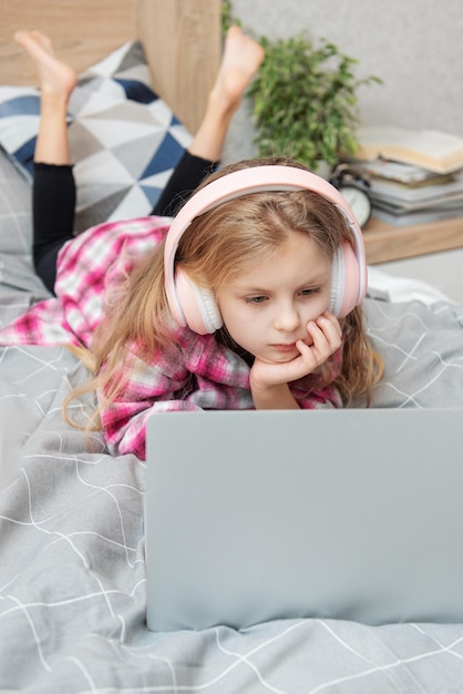 Glimlachend gelukkig klein meisje met laptop in draadloze koptelefoon in bed thuis