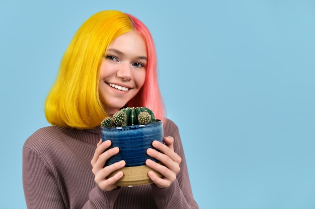 Glimlachend gelukkig tienermeisje hoofd geschoten met geverfd gekleurd haar met pot met cactus kamerplant, op blauwe studio achtergrond