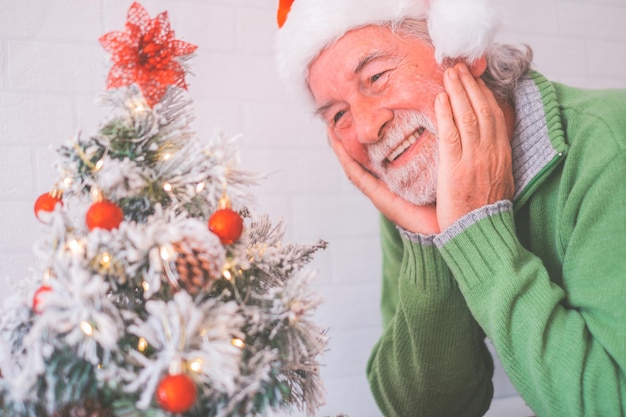 Glimlachend gepensioneerde senior man in kerstmuts en warme kleding kerstboom thuis versieren. Oude mannelijke kerstman glimlacht terwijl hij naar een versierde kerstboom kijkt. Oudere man geniet van kerstvakantie