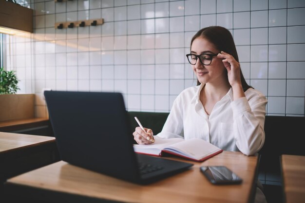 glimlachend hipstermeisje in wit overhemd dat huiswerk doet om informatie te controleren via moderne laptopcomputer