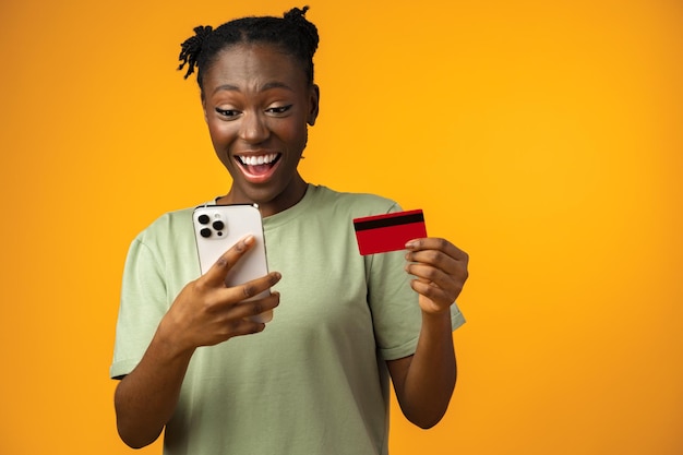 Glimlachend jong afro-meisje houdt smartphone en creditcard vast in gele studio