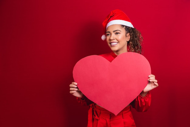 Glimlachend jong afro-meisje in kerstmuts met een enorme hartkaart met vrije ruimte voor tekst.