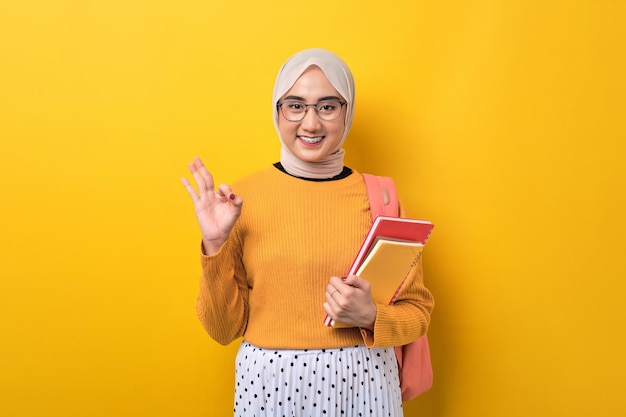 Glimlachend jong Aziatisch studentenmeisje met het notitieboekje van de rugzakholding die ok teken met vinger doen die over gele achtergrond wordt geïsoleerd