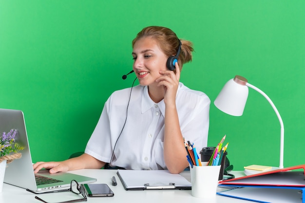 Glimlachend jong blond callcentermeisje met een hoofdtelefoon die aan een bureau zit met uitrustingsstukken die de hoofdtelefoon aanraken met een laptop die op een groene muur is geïsoleerd