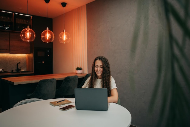 Glimlachend jong brunette meisje met lang golvend haar in een beige T-shirt werkt thuis achter een laptop, een notitieboekje met een pen en een smartphone ligt op tafel