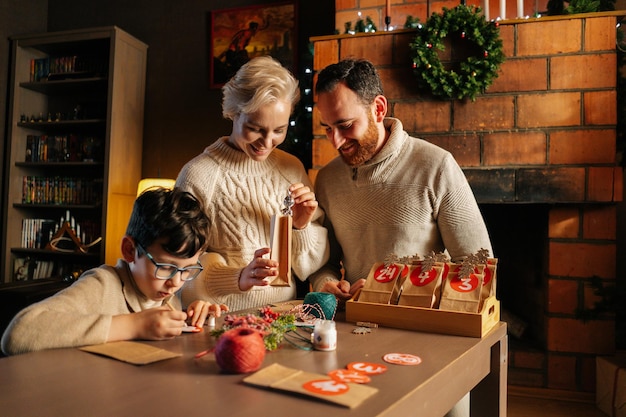 Glimlachend jong gezin maken kerst adventkalender samen zittend aan tafel op de achtergrond van de open haard in een gezellig huis op kerstavond