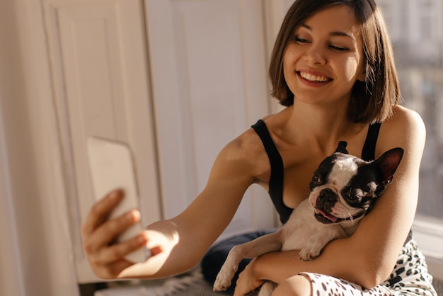 Glimlachend jong kaukasisch meisje neemt een foto van zichzelf op de telefoon en houdt de hond bij het raam Love for pets joy and technology