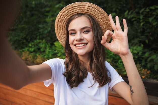 Glimlachend jong meisje dat een selfie met uitgestrekte hand neemt in het park buitenshuis, ok weergegeven