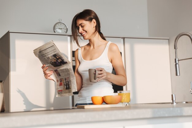 Glimlachend jong meisje dat in de ochtend bij de keuken staat, krant leest, thee drinkt