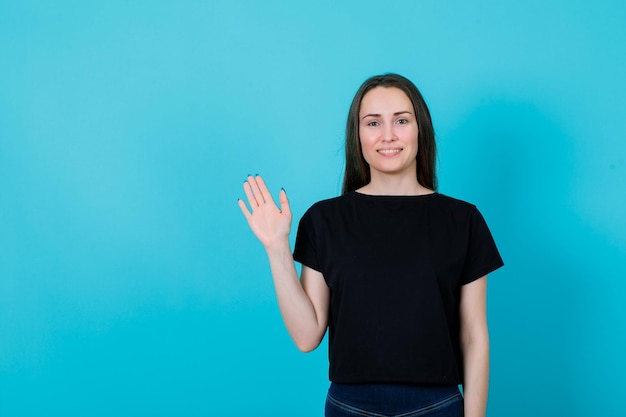 Glimlachend jong meisje toont hallo gebaar door haar hand op te steken op een blauwe achtergrond