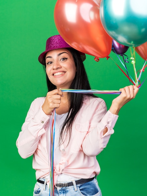 Glimlachend jong mooi meisje met feestmuts met ballonnen geïsoleerd op groene muur