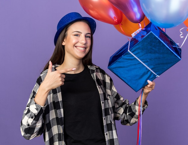 Glimlachend jong mooi meisje met feestmuts met ballonnen met geschenkdoos geïsoleerd op blauwe muur