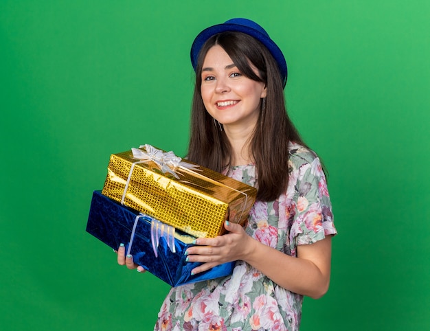 Glimlachend jong mooi meisje met feestmuts met geschenkdozen geïsoleerd op groene muur