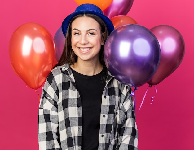 Glimlachend jong mooi meisje met feestmuts staande voor ballonnen geïsoleerd op roze muur