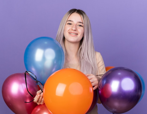 Glimlachend jong mooi meisje met tandheelkundige beugels die achter ballonnen staan met een bril geïsoleerd op een blauwe muur