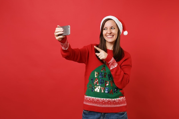 Glimlachend jong Santa meisje doen selfie schot op mobiele telefoon, wijzende wijsvinger geïsoleerd op heldere rode achtergrond. Gelukkig Nieuwjaar 2019 viering vakantie partij concept. Bespotten kopie ruimte.