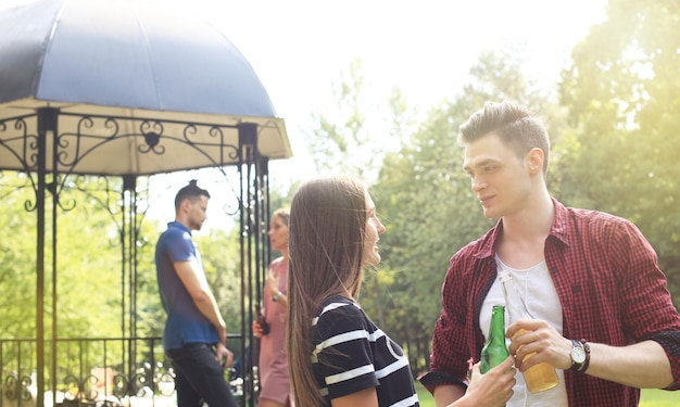 Glimlachend jong stel met bier en kijken naar elkaar terwijl twee mensen barbecueën op de achtergrond.