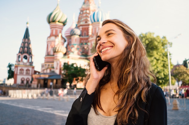 Glimlachend jong vrouwenportret in Rood Vierkant die bij de telefoon, met de Kathedraal van het Basilicum van Heilige op de achtergrond spreken. Moskou, Rusland.
