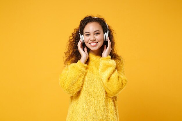 Glimlachend jonge Afro-Amerikaanse meisje in bont trui poseren geïsoleerd op geel oranje achtergrond in studio. Mensen oprechte emoties levensstijl concept. Bespotten kopie ruimte. Luister muziek met een koptelefoon.