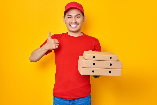 Glimlachend jonge Aziatische man in rode dop tshirt uniforme levering werknemer werk als dealer koerier houden pizzadoos van restaurant duim omhoog teken geïsoleerd op gele achtergrond Pizza levering concept