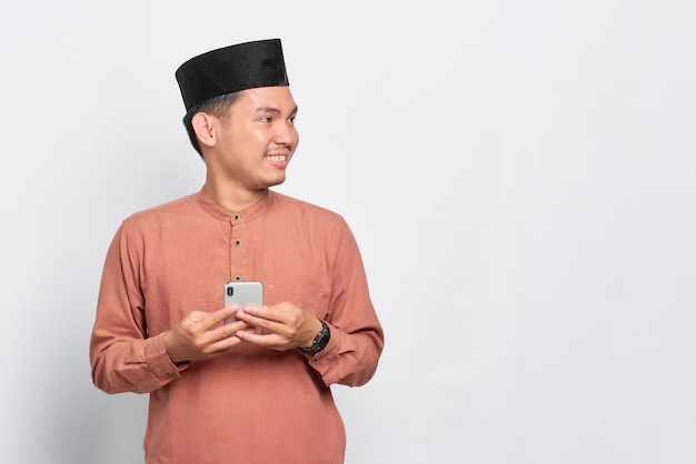 Glimlachend jonge Aziatische moslim man met behulp van een mobiele telefoon en wegkijken geïsoleerd op witte achtergrond