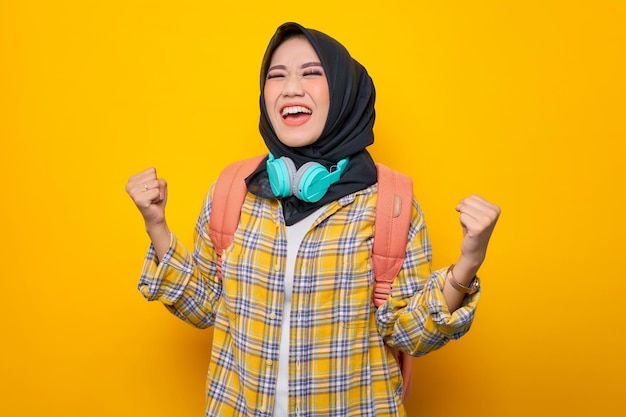 Glimlachend jonge Aziatische moslimvrouw student in plaid shirt met koptelefoon en rugzak met behulp van mobiele telefoon en winnaar gebaar op gele achtergrond Onderwijs school universiteit college concept