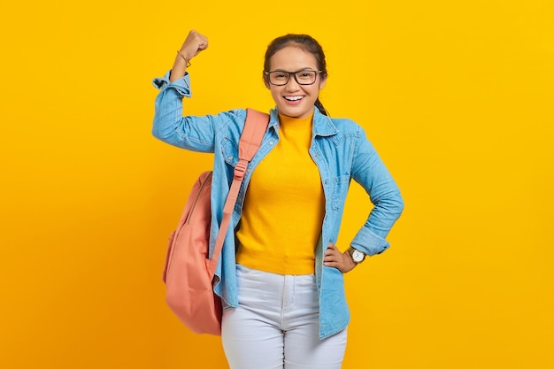 Foto glimlachend jonge aziatische student in denim outfit met rugzak handen opsteken en tonen van biceps prestaties geïsoleerd op gele achtergrond onderwijs in middelbare school universiteit college concept