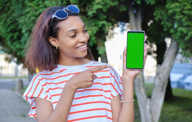 Glimlachend jonge zwarte vrouw wijst naar haar mobiele telefoon scherm mobiele telefoon scherm in groene kleur voor bijsnijden draagt ze een beige gekleurde blouse geïsoleerd op beige achtergrond