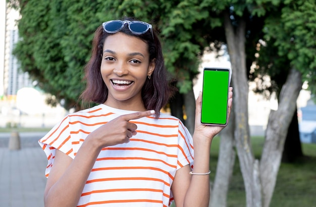 Glimlachend jonge zwarte vrouw wijst naar haar mobiele telefoon scherm mobiele telefoon scherm in groene kleur voor bijsnijden draagt ze een beige gekleurde blouse geïsoleerd op beige achtergrond