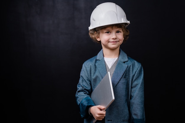 Glimlachend jongetje in bouwvakker en formalwear die gevouwen laptop houdt terwijl hij geïsoleerd tegen zwarte ruimte staat